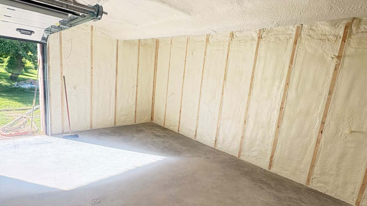 Garaazi soojustamine 1 Unfinished garage interior with spray foam insulated walls exposed vertical studs and a concrete floor open garage door shows a grassy yard outside