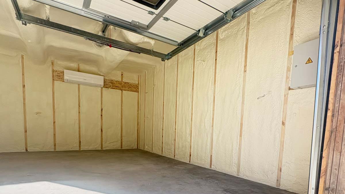 Garaazi soojustamine 2 Empty garage interior with spray foam insulation exposed wooden studs and overhead garage door rails The concrete floor is bare